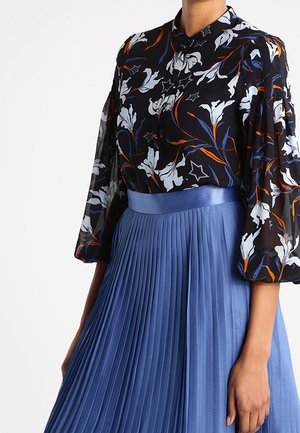 Mujer con una blusa negra floral con mangas abullonadas metida en una falda azul plisada de talle alto.