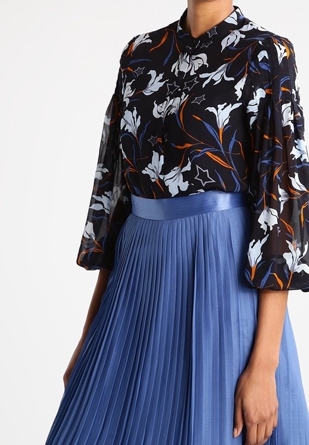 Femme portant un chemisier noir fleuri avec des manches bouffantes, rentré dans une jupe plissée bleue taille haute.