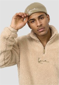 Beige színű polárpulóver sekély zipzárral, texturált anyaggal és arany színű logóval. A modell fején egy szintén beige színű kötött sapka.