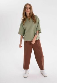 Överdimensionerad olivgrön sweatshirt med en framficka, kombinerad med bruna vida byxor och vita sneakers, mot en vit bakgrund.