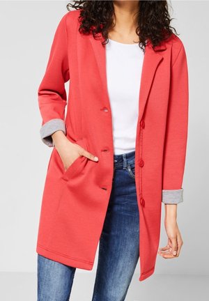 Kobieta nosząca koralową długą marynarkę z podwiniętymi szarymi mankietami, na białej bluzce i niebieskich dżinsach, jedną ręką w kieszeni, na jednolitym tle.