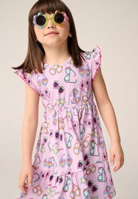 Růžové šaty s krátkými nabíranými rukávy, zdobené barevným vzorem brýlí, květin a ovoce. Dítě má na sobě kulaté, vícebarevné sluneční brýle.