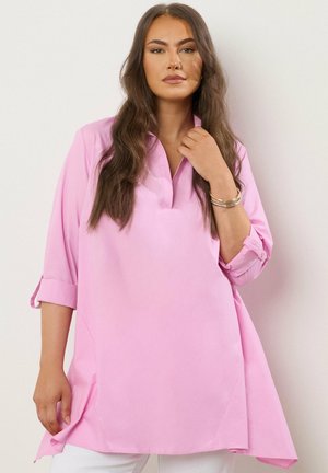 Kobieta o długich brązowych włosach, ubrana w luźną różową koszulę z podwiniętymi rękawami i białe spodnie, stojąca na prostym tle.