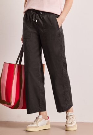 Personne portant un pantalon noir à cordon, des baskets blanches avec des accents beige et rose, tenant un grand sac fourre-tout rayé rouge et rose.
