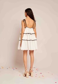 Vestido branco em camadas com detalhes em preto, costas abertas e alças spaghetti ajustáveis. Usado com sandálias de salto bloco em bege. Confete colorido espalhado.