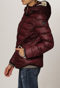 Blouson rembourré bourgogne avec capuche, présentant un design matelassé, des poches zippées et une doublure texturée. Mi-long avec des manches ajustées.