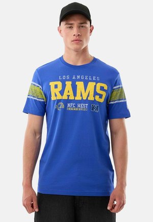 Jonge man draagt een blauw Los Angeles Rams T-shirt met gele tekst en een zwarte honkbalpet, staand tegen een effen achtergrond.