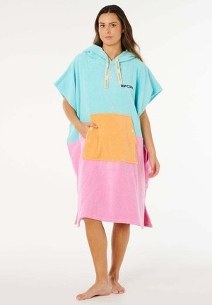 Kvinde iført en løs, hætteklædt ponchohåndklæde i blå, pink og orange med "Rip Curl"-logo, stående barfodet mod en hvid baggrund.