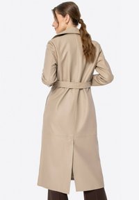 Beige trench coat vyrobený z hladkého materiálu, s opaskem v pase, velkým límcem a středovým rozparkem na zadní straně pro snadný pohyb.