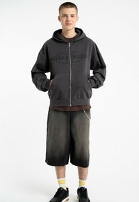 Szara bluza z kapturem zapinana na zamek z dużym napisem "INFLAMMABLE". Połączona z ciemnoniebieskimi szortami do kolan i czarnymi trampkami z żółtymi paskami.