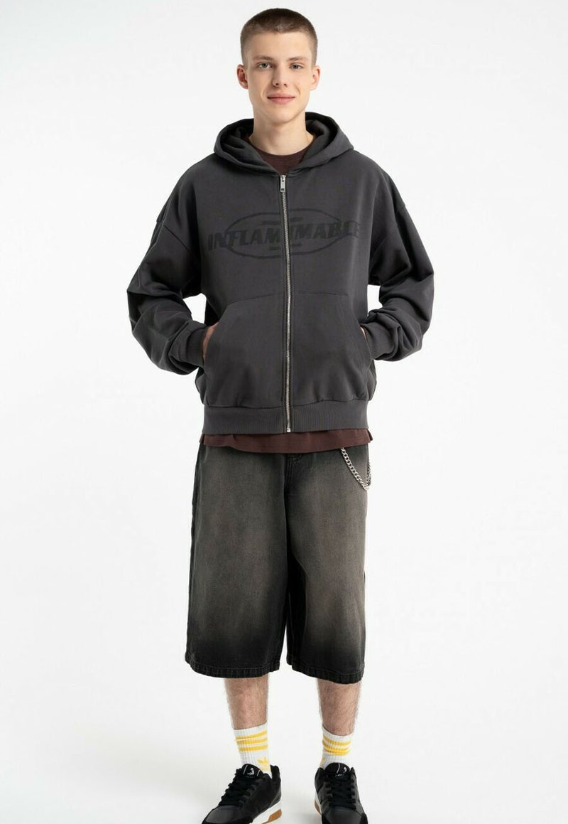 Szara bluza z kapturem zapinana na zamek z dużym napisem "INFLAMMABLE". Połączona z ciemnoniebieskimi szortami do kolan i czarnymi trampkami z żółtymi paskami.
