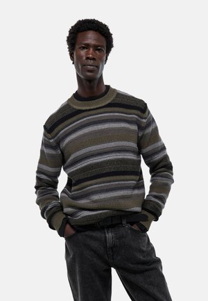 Pull en maille rayé en olive, gris et noir avec un col rond ; motif texturé ; porté avec un jean foncé ; coupe décontractée.