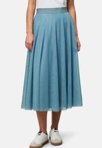 Une longue jupe plissée en tulle bleu clair avec une taille lisse, associée à des baskets blanches. Le tissu reflète la lumière en douceur.