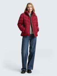 Rote gesteppte Daunenjacke mit hohem Kragen und Reißverschluss, getragen über einem grauen Pullover, kombiniert mit weit geschnittenen Jeans und schwarzen Schuhen.