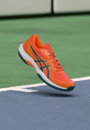 Lyse orange sportssko med sorte striber og hvid sål svæver over en blå og grøn tennisbane nær grænselinjen.