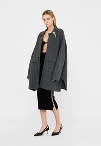 Grijze oversized jas van een wolmix, met een knoopsluiting aan de voorkant, grote zakken en een asymmetrische zoom, gecombineerd met een zwarte kokerrok.