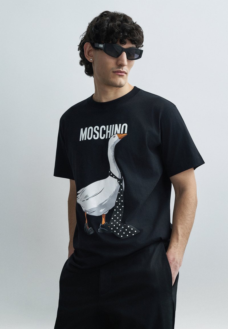 Čierne bavlnené tričko s grafikou bielej husi v kravate s bodkami a topánkach. Názov značky "Moschino" je zobrazený nad tým.