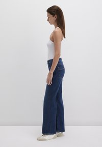 High-waisted, dark blue denim jeans with a wide leg design, featuring back pockets and stitching details. Worn with a white tank top and white shoes.
