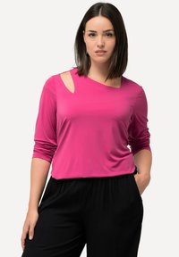 Top fucsia a maniche lunghe con un'apertura sulla spalla sinistra, realizzato in un tessuto liscio e leggero, abbinato a pantaloni neri plissettati.