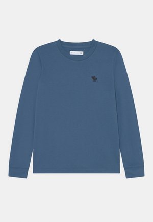 Camiseta azul de manga larga con cuello redondo y pequeño logo de alce negro en el lado izquierdo del pecho, mostrada sobre un fondo neutro.