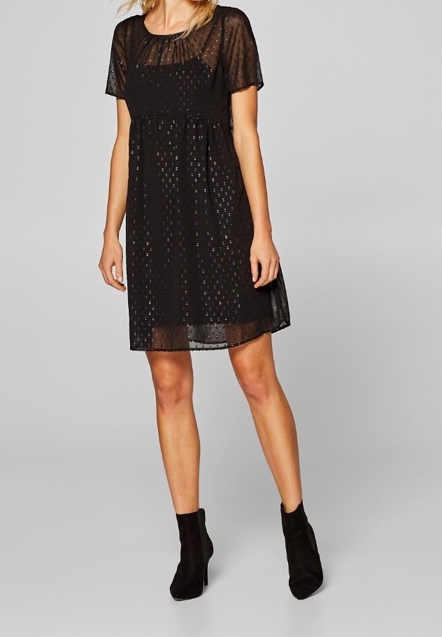 Femme portant une robe courte noire avec un voile transparent à pois et des manches courtes, associée à des bottines noires.
