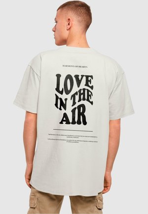 T-shirt à manches courtes en coton crème avec un texte graphique noir sur le dos indiquant « LOVE IN THE AIR », accompagné d'un texte plus petit au-dessus.