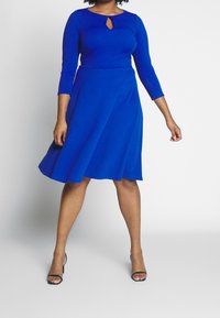 Une robe bleue longueur genou avec un corsage ajusté, des manches trois-quarts et un détail d'encolure en forme de trou de serrure ; fabriquée en tissu doux et extensible.