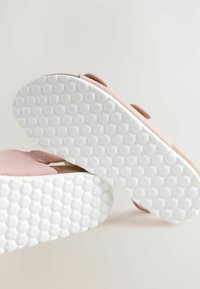 Lichtroze sandalen hebben een zachte suede bovenkant met klittenbandsluitingen en een gestructureerde witte rubberen zool met een honingraattracering.