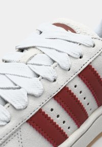 Zapatilla blanca con rayas de cuero rojo, parte superior texturizada, cordones blancos planos y una suela de goma contrastante con un patrón sutil.