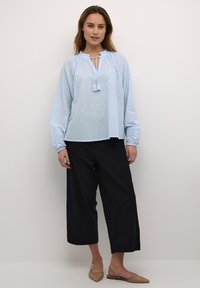 Lichtblauwe blouse met lange mouwen en een trekkoord in de hals, versierd met franjes. De blouse heeft een broderie anglaise-detail bij de manchetten en is gecombineerd met zwarte wijde broek.