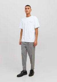 Camiseta de algodón blanca de manga corta, con un pequeño gráfico de texto negro, combinada con pantalones ajustados grises claros y zapatos negros.