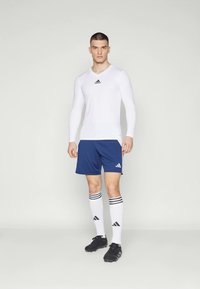 Balta sporta krekls ar garām piedurknēm un melnu logotipu, kombinēts ar tumši zilām šortiem, baltiem pusceļa augstiem zeķēm ar melnām svītrām un melnām futbola kedām.