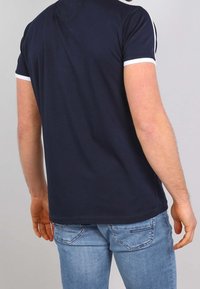 Námořnické modré tričko s krátkým rukávem a bílým límcem a bočními akcenty, které má uvolněný střih a hladkou bavlněnou texturu. Nosené s světle modrými džínami.