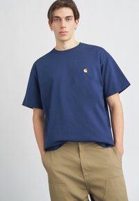 Temno modra bombažna majica z ohlapnim krojem, okroglo izrezano ovratnikom in majhnim rumenim logotipom na levem prsnem delu. Nosimo jo z bež hlačami.