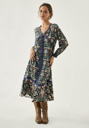 Mujer con un vestido largo y estampado con diseños florales en azul y beige, y botas de ante marrones, de pie frente a un fondo claro y liso.