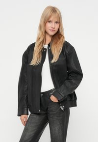 Blonde vrouw in een zwart leren jasje over een witte top en zwarte gebreide broek, staande met één hand in de zak tegen een witte achtergrond.