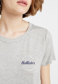 Šedé tričko s krátkým rukávem, kulatý výstřih, s logem "Hollister" v námořnické modři a kulatým designem s textem "Southern California". Měkká textilie.