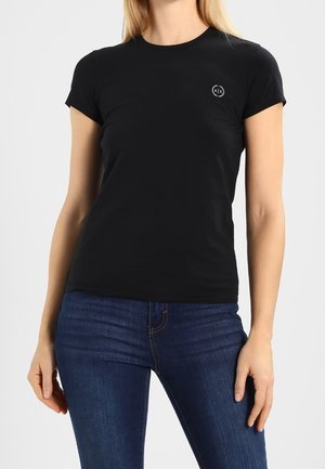 Kobieta nosząca dopasowany czarny t-shirt z krótkim rękawem z małym okrągłym logo na piersi oraz niebieskie dżinsy.