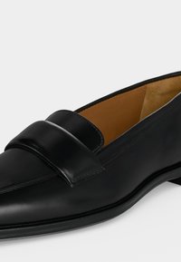 Μαύρο δερμάτινο loafer με μυτερή μύτη και λεπτομέρεια με λουρί, ταμπά εσωτερική επένδυση και χαμηλό τακούνι σε λευκό φόντο.