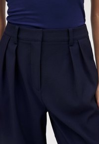 Tumed sinise värvi kohandatud püksid, millel on voldid ees ja küljetaskud, valmistatud siledast kangast, millel on struktureeritud istuvus.