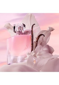 Klar glass parfymeflaske med en rosa væske og et glitrende rosa bånd rundt halsen, prydet med en delikat sløyfe og orkidéblader.