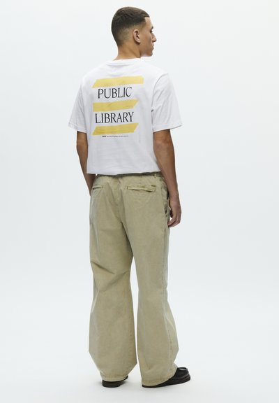 Oldalra néző férfi, fehér "KÖNYVTÁR" feliratú pólóban és széles bézs nadrágban, egyszínű háttér előtt.