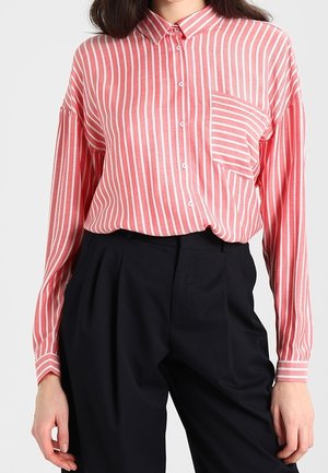 Vrouw draagt een lange mouwen roze-wit verticale gestreepte blouse, ingestopt in hoog getailleerde zwarte broek.