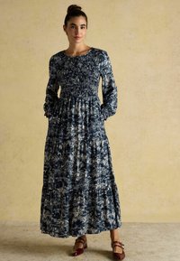 Maxi šaty z tmavomodrej látky s bielymi kvetinovými vzormi, dlhými rukávmi, elastickým telom, volánovou sukňou a bočnými vreckami. Hnedé topánky.