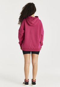 Mujer con cabello largo y rizado, vistiendo una sudadera holgada magenta, pantalones cortos de ciclista negros y zapatillas altas negras, de pie de espaldas.