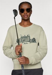 Ανοιχτό πράσινο φούτερ με σκούρα γραφικά ενός κάστρου και το κείμενο "Malbon". Το μοντέλο φορά γυαλιά ηλίου και κρατά ένα γκολφ κλαμπ, με μαύρη λαβή.