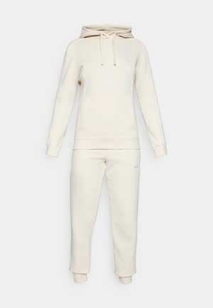 Kreminės spalvos Puma mikrofonas su raišteliais ir atitinkančios jogger kelnės, abiejuose yra subtilūs Puma logotipai ant krūtinės ir šlaunies.
