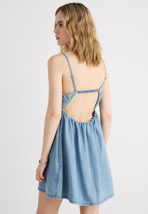 Frau mit schulterlangem, welligem blondem Haar, die ein ärmelloses hellblaues Jeanskleid mit offenem Rücken und dünnen, verstellbaren Trägern trägt.