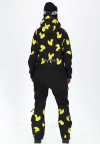 Skikostuum van zwart flexibel materiaal met gele rubberen eendjespatronen. Inclusief een capuchon en ritsen op de broek voor verstelbaarheid.