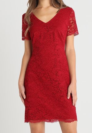 Robe courte à manches courtes en dentelle rouge avec un décolleté en V, motif floral et ourlet festonné, portée par une femme debout devant un fond uni.